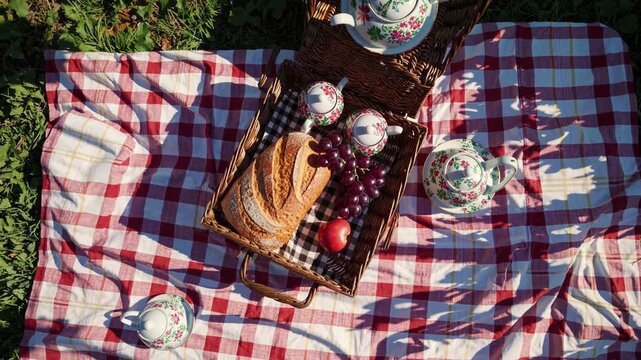 Bread rests in woven basket with grapes, apple, and delicate floral cups. Red checked blanket spreads across grass, creating warm picnic setting under crisp daylight with sliced loaf and tableware.
