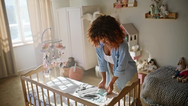 Expectant mother laying baby clothes crib. Happy woman touching belly nursery.