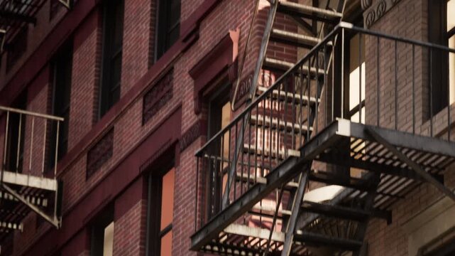 Rustic brick buildings stand tall against the sky, showcasing characteristic fire escapes. The lively scene captures the essence of city life and architectural history during the day.