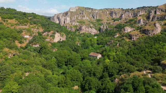 Aerial drone footage flying over a forested canyon toward mountain cliffs with visible caves and rock formations in southern Armenia.