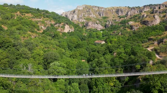 Aerial drone footage ascending from a suspension bridge over a canyon toward higher mountain terrain, revealing a green gorge, steep rock cliffs and cave formations in southern Armenia.