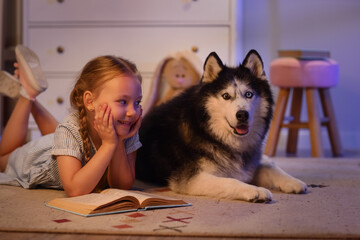 Childhood companionship and reading, little girl lying on floor with her husky dog at home during...