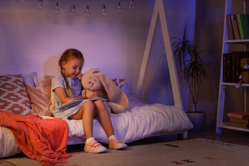Childhood education and bedtime reading, little girl reading a book with a plush toy in a cozy bedroom at night with warm string lights © Pixel-Shot