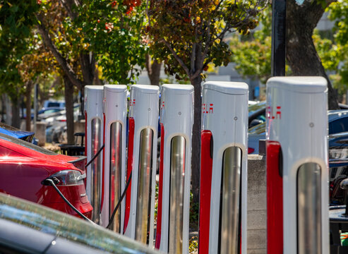 Outdoor Tesla charging area featuring white‑and‑red Supercharger stations and multiple parked electric vehicles in everyday use