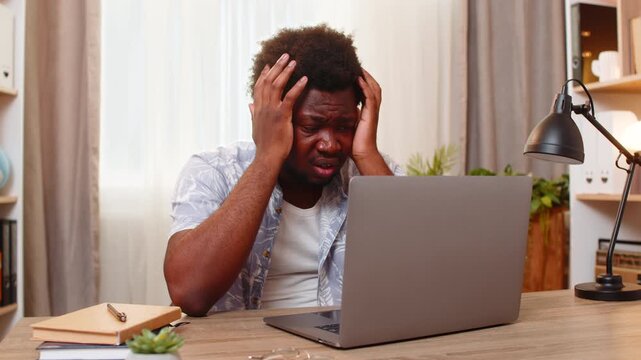 African American young man on home table stares at laptop in shock after reading bad news online and holds head from stress. Black guy on home table feels upset and helpless after an alarming message.