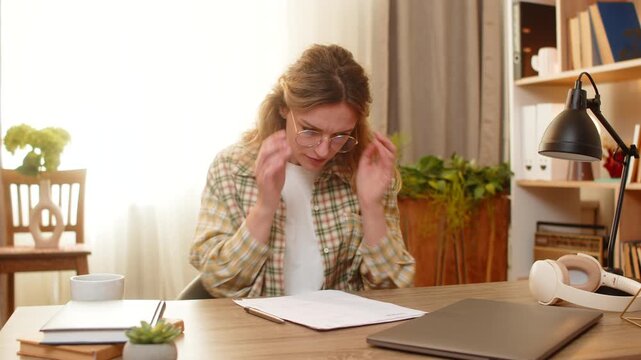 Young woman on home table puts on glasses reads fine print in document and writes note with pen during careful review. Adult girl on home table checks paper details with focused frown for remote work.