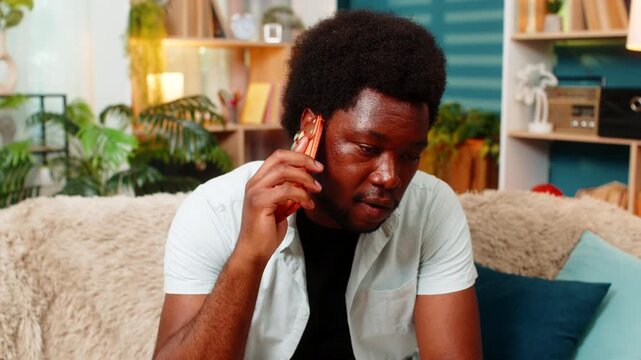 African American young man on home sofa answers smartphone call and reacts with tired face during unwanted talk indoors. Black guy on home sofa speaks on phone and feels stressed by annoying caller.