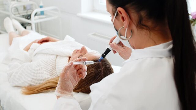 Caucasian girl undergoes scalp treatment to stimulate hair regrowth using microneedling dermapen and vitamin injections. Procedure performed by trichologist in cosmetology clinic for hair restoration.