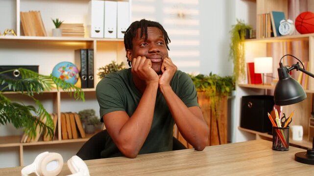 African American young man on home table rests on hands and daydreams with gentle smile during peaceful break. Black guy on home table looks upward imagining future plans in cozy living room indoors.