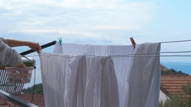 Woman hanging fresh white laundry on clothesline. Housewife hanging clean white laundry on a clothesline on the balcony with a beautiful sea view in the background, completing a household chore