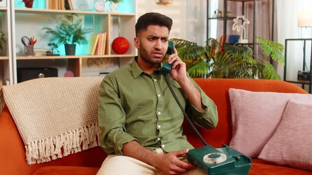 Indian man on home sofa listens to old wired phone with shocked worried expression after strange call. Arabian guy on home sofa feels stress during retro telephone talk about urgent home news indoors