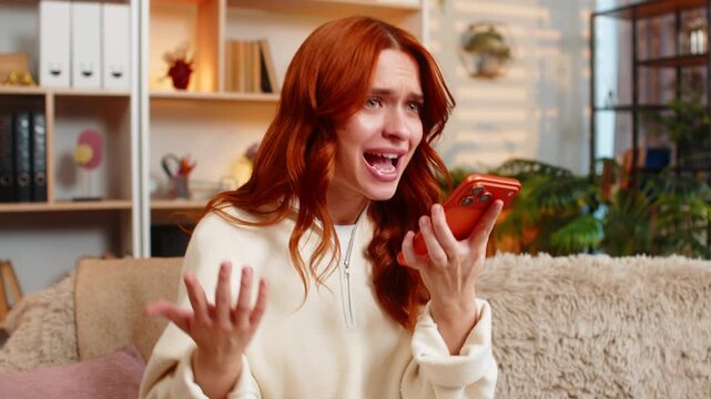 Young woman on home sofa talks on smartphone speaker and waves hand in furious dispute showing frustration. Adult girl on home sofa reacts with outrage during heated call and voices strong complaint.