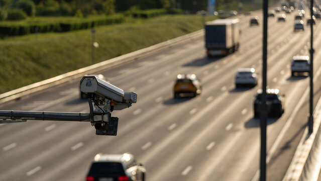 Traffic camera monitors a busy highway. Cars travel on multiple lanes under bright sky. Grassy field borders the roadside. Ideal for traffic safety or security projects