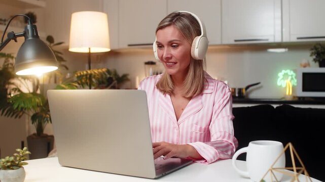 Adult woman at night home edits layout on laptop in headphones listens to music smiles nods. Caucasian girl at table on sofa at evening checks preview applies changes saves file enjoys rhythm steady