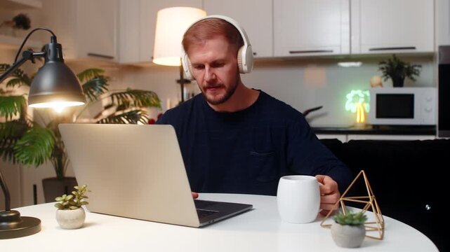 Young man at night home opens laptop does freelance work drinks tea wears wireless headphones listens to music focused. Caucasian guy on sofa at table types on pc does remote job productive indoors.