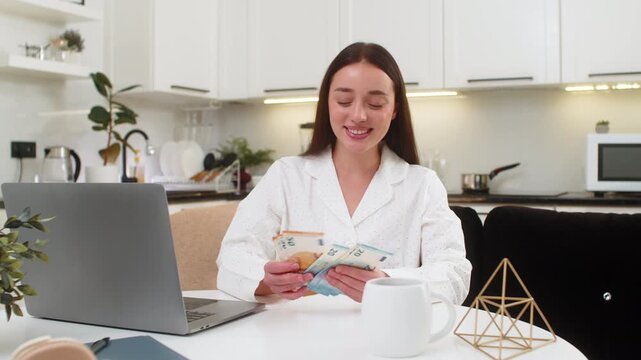 Young woman at home counts money cash euros smiles feels proud about salary, savings plans to invest. Caucasian girl on sofa sits at table stacks bills budget track finance calculates amount and goals
