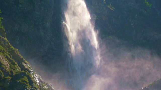 Waterfall cascades through rocky Alps landscape. Waterfall flows down alpine mountain cliff. Mountain waterfall splashes over mossy rocks. Natural stream flows below waterfall.