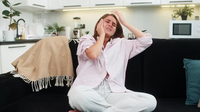 Young woman at home feels toothache holds cheek and presses palm to jaw with short wince and pain. Caucasian girl on sofa breathes slow rubs jaw and looks for relief and rest before dentist visit fast