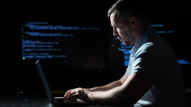 Programmer coding on laptop in dark room. Programmer typing beside blue source code. Focused programmer working with computer technology. Male programmer at illuminated digital workspace.