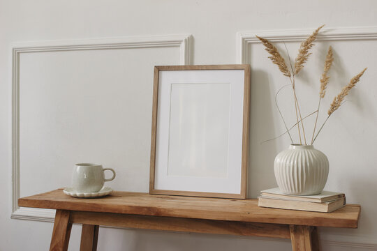 Minimalist Scandinavian interior. Blank framed poster, picture frame mockup on wooden console table. Ceramic vase with dried grasses, coffee cup, books. Neutral beige home decor, copy space. Side view