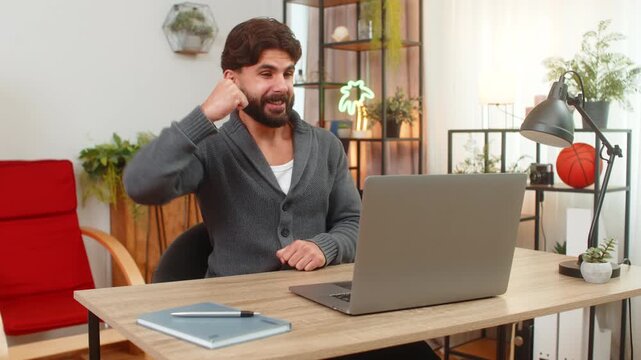 Indian man at home reads email on laptop partnership confirmed smiles raises thumb celebrates news. Arabian guy at desk nods approval feels relief success after long wait positive message brings joy