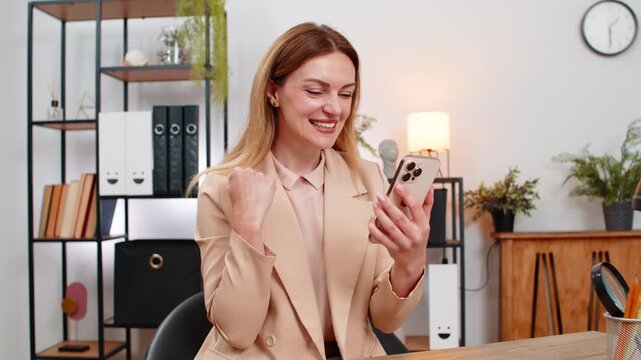 Middle-aged businesswoman at home office taps app on smartphone wins promo discount smiles lifts fist and hears news of salary boost. Freelancer girl at table confirms win applies code and saves big