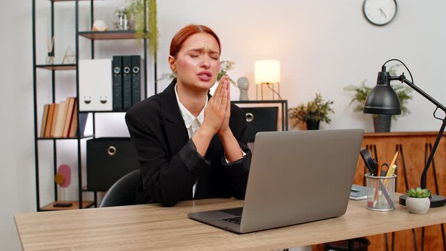 Young businesswoman at home office clasps hands before laptop looking up praying for project approval. Freelancer girl by table hopes order arrives trusting good faith and thinking to secure success.