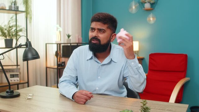 Indian man at home shakes empty piggy bank unhappy debt weighs shows bankruptcy poverty big despair. Arabian guy at desk sighs sadness grows as coins absent financial loss frustration worry over money