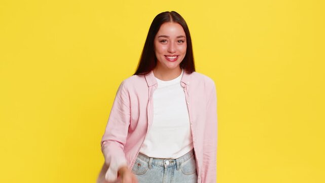 Young woman smiling gently and forming heart shape with fingers, expressing love kindness and affection through warm gesture. Girl on yellow background shows emotional connection and positive feelings
