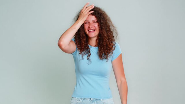 Young woman making facepalm gesture covering face with hand, laughing humorously at silly, funny mistake. Girl on gray background expresses humorous reaction to embarrassing misunderstanding situation