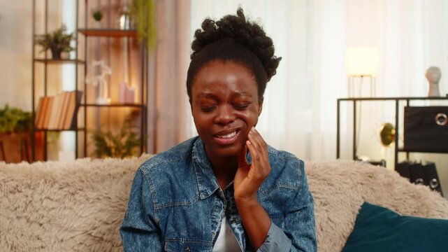 African American young woman painfully touches her cheek and closes her eyes from a sharp toothache, looking visibly unwell and distressed. Black girl suffers dental pain while sitting at home.