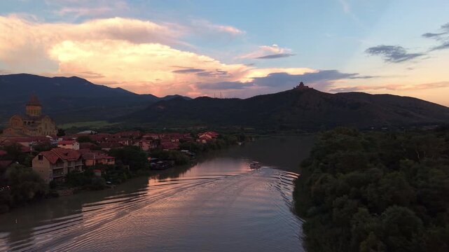 Aerial sunset view of a boat crossing the Mtkvari River in historic Mtskheta, Georgia, framed by Svetitskhoveli Cathedral, mountain ridges and a glowing evening sky.