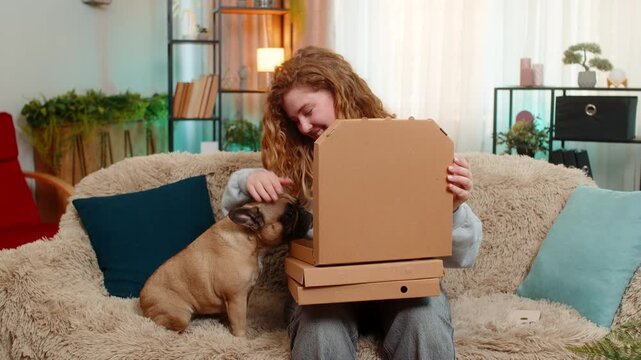 Young Caucasian redhead woman opens pizza boxes on home sofa, smiling at delicious meal with excitement. Pug dog bites slice playfully, sharing happy moment with owner during food delivery treat.