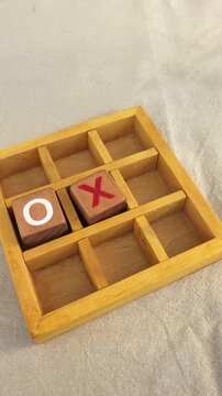 Two players take turns using wooden pieces to play tic-tac-toe on a small game board. The action shows the placement of moves on a light surface.