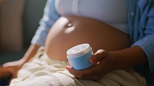 Closeup hands applying moisturizer cream pregnant belly. Skincare home sofa.