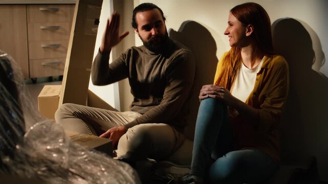 Proud partners sharing a high five after moving into their new house, sitting on the floor and discussing furniture placement and interior design layout.