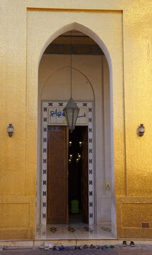 A pointed arch entrance adorned with gold mosaic tiles, a hanging metal lantern, and shoes lined up outside leads into the Golden Mosque at Katara Cultural Village in Doha, Qatar