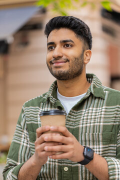 Indian man drinking coffee walking in city street with calm smile enjoying morning stroll and travel mood. Arabian guy tourist holding takeout cup outdoors looking at buildings during calm urban walk.