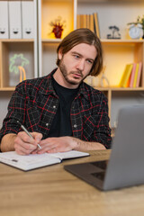 Fototapeta premium Young man on home table watches study video on laptop stops playback and writes ideas in notebook. Adult guy on home table stays focused on online learning and keeps taking notes with calm attention.