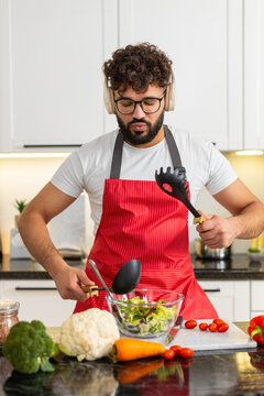 Arabian man dances with headphones while mixing fresh salad in kitchen for healthy diet and clean eating. Young guy enjoys music and cooks fresh vegetables at home apartment feeling joyful fun calm.