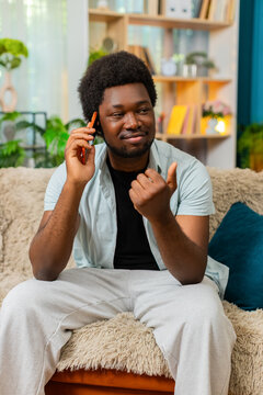 African American young man on home sofa answers smartphone call with calm serious face during private talk at home. Black guy on home sofa listens to mobile phone news for work or family talk indoors.