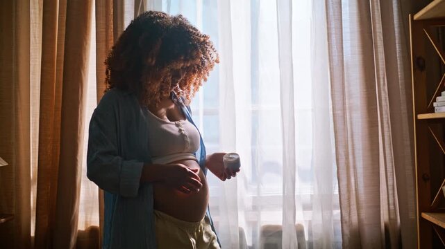 Pregnant woman applying belly cream window. Maternity skincare stretch marks.