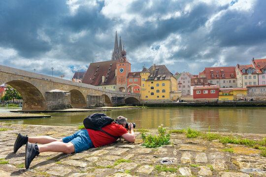 Photographer taking pictures beside the Danube River with the Stone Bridge and historic architecture of Regensburg, Bavaria, Germany.