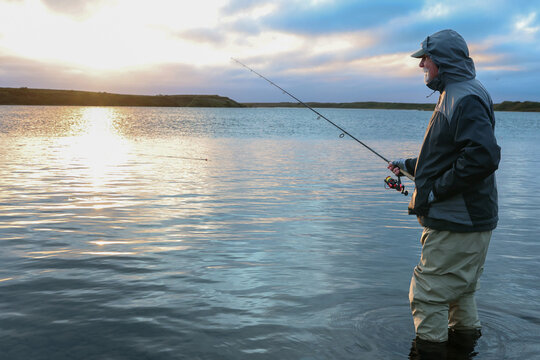A mature man wearing waders and a rain coat holds a fishing pole while salmon fishing on the Egegik River in Alaska during a beautiful, quiet sunrise, surrounded by calm water and soft morning light.