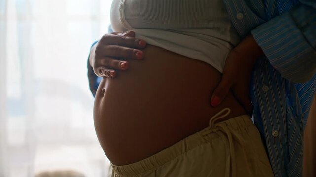 Expectant mother stroking baby bump closeup. Pregnant belly hands window light.