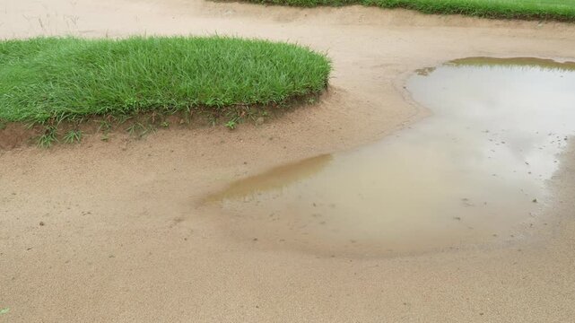 The camera pans right over a sand trap on a wet golf course, due to rain.