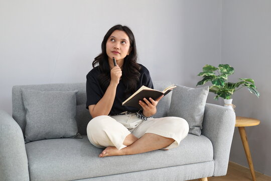 A young woman is focused on writing in a notebook while sitting on a comfortable sofa; she appears to be deep in thought and reminiscing, radiating creativity and inspiration as she looks upward.