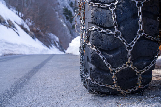A large vehicle tire fitted with metal snow chains stands on a paved mountain road beside melting snowbanks, a concept of winter driving safety.