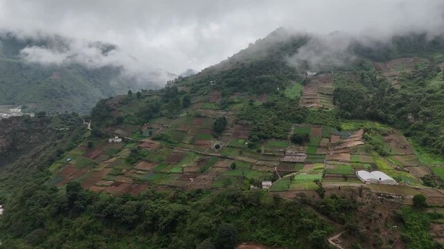 Drone flies forward over patchwork vegetable farms on green mountain slopes as beautiful low clouds drift across Zunil, Quetzaltenango, Guatemala.