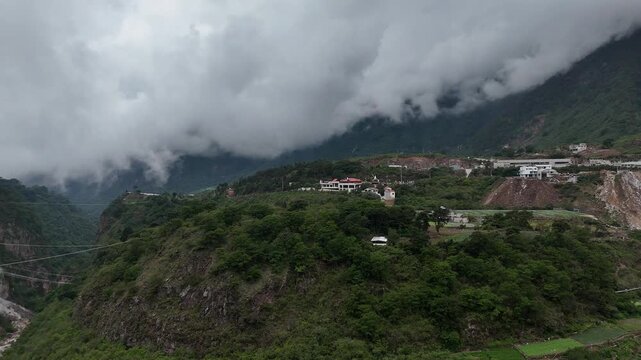 Drone flies forward toward a rural mountain settlement surrounded by steep green slopes and dramatic low clouds in Zunil, Quetzaltenango, Guatemala.